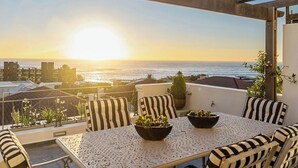 A balcony with a table and chairs, overlooking a sunset over the ocean. - Luxury villa 120m from beach & restaurants striking ocean & mountain view-by JGM (Cape Town)