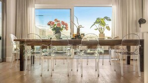A dining area with a wooden table and clear chairs, large windows with curtains, and potted plants. - Luxury villa 120m from beach & restaurants striking ocean & mountain view-by JGM (Cape Town)