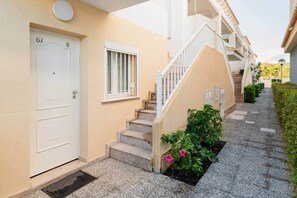 A residential building with a beige exterior, a white door labeled '67', a window with white bars, a staircase with white railings, and a small garden with pink flowers. - House with Barbecue Pool and Private Parking - Zona norte playa Gandía (Gandia)