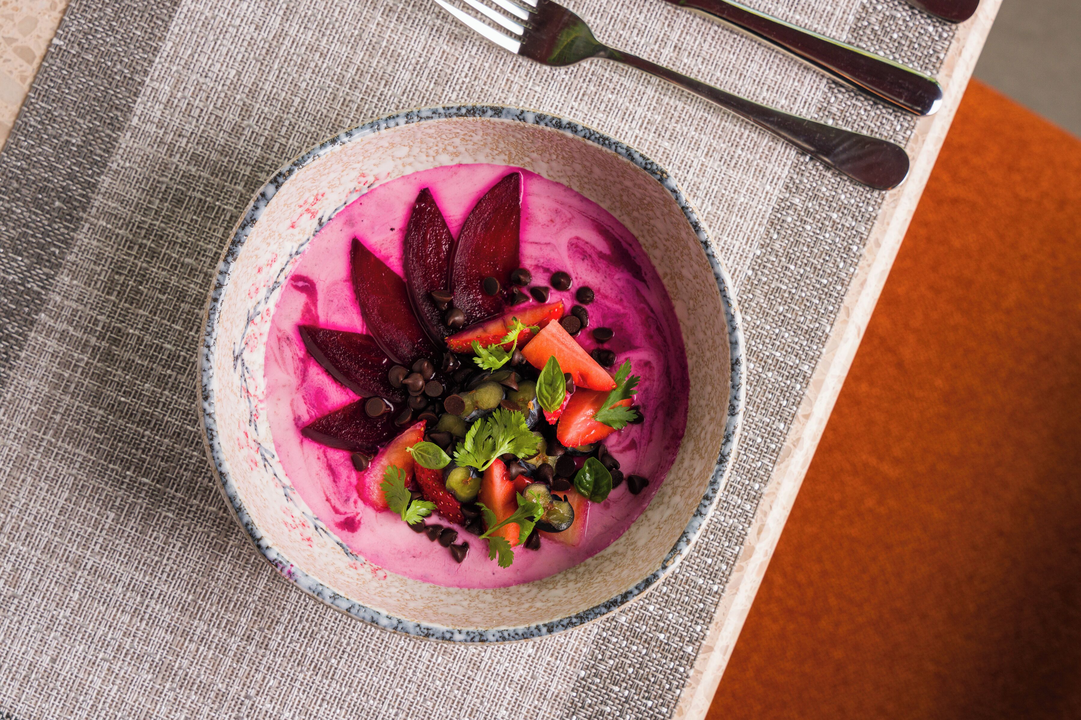Ein farbenfroher, violetter Smoothie mit Obst und Schokoladenstückchen auf einer strukturierten Tischdecke.