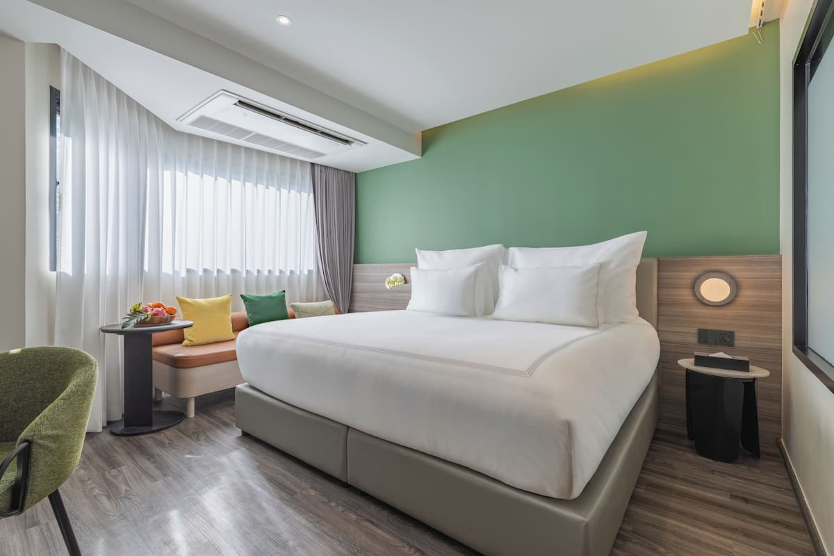 A modern hotel room with a large bed, a small side table, a chair, and a window with curtains.