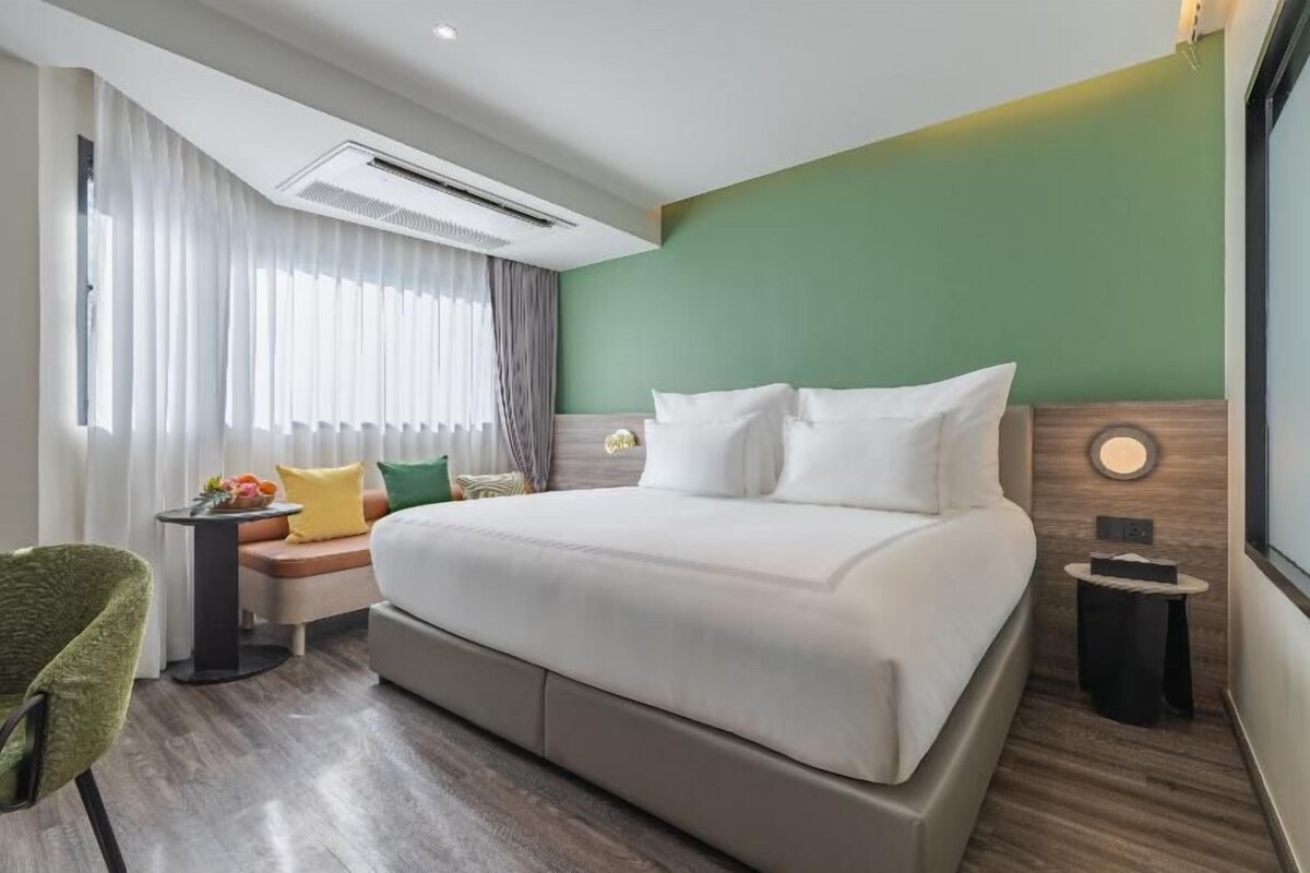 Ein Hotelzimmer mit einem großen Bett, einem Nachttisch, einem Sessel und einem Fenster mit Vorhängen.