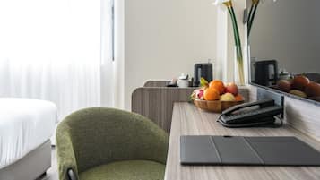 A hotel room with a bed, a green armchair, a wooden desk with a fruit bowl, a telephone, and a mirror reflecting the room.