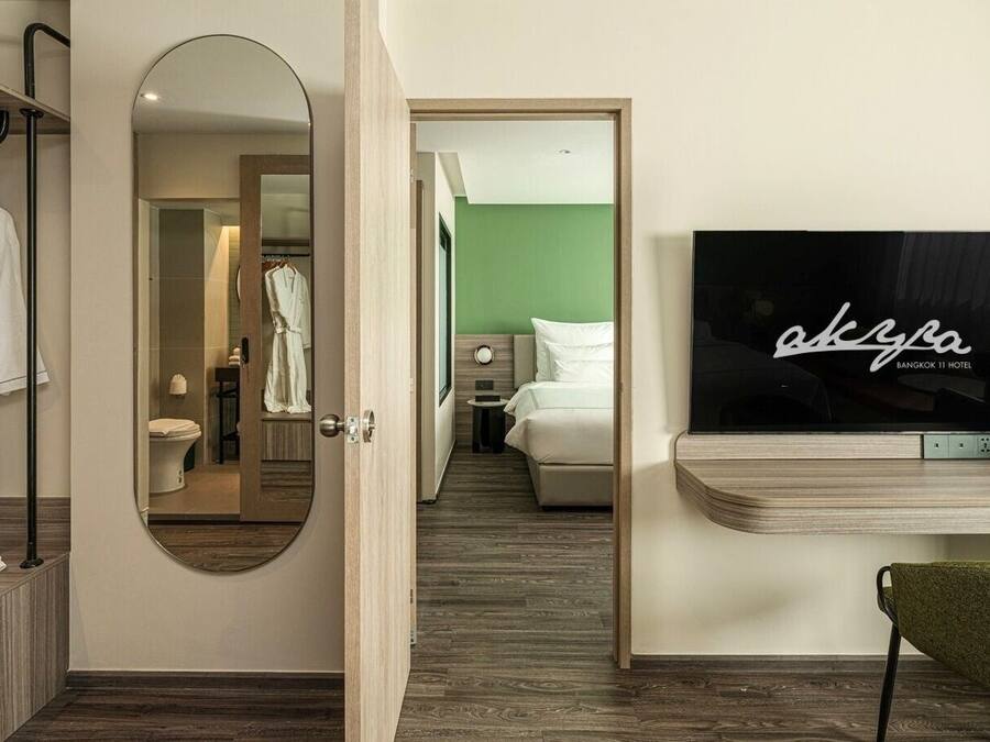 Een hotelkamer met een houten opbergmeubel, een spiegel, een groene stoel, een bed en een televisie waarop de naam van het hotel staat.