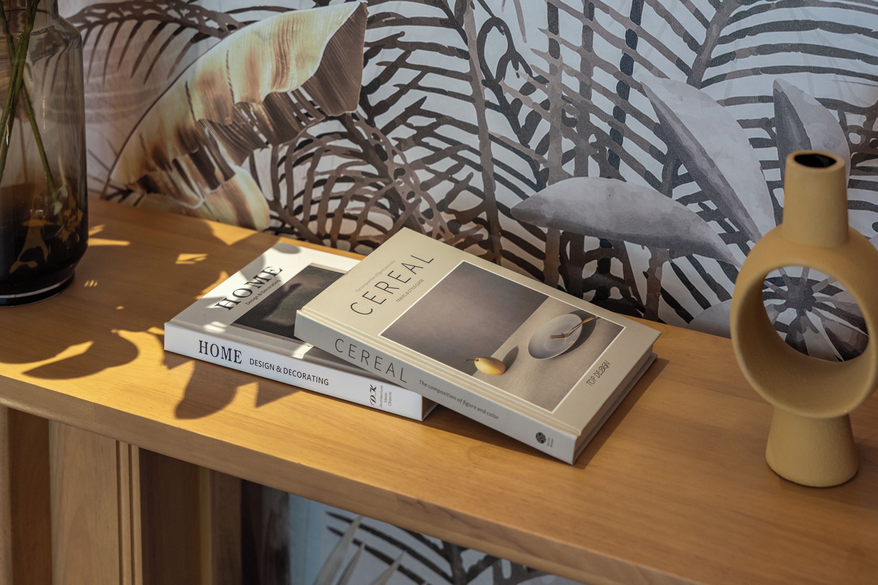 A wooden table with books titled 'HOME DESIGN & DECORATING' and 'CEREAL,' a vase, and a patterned wall.