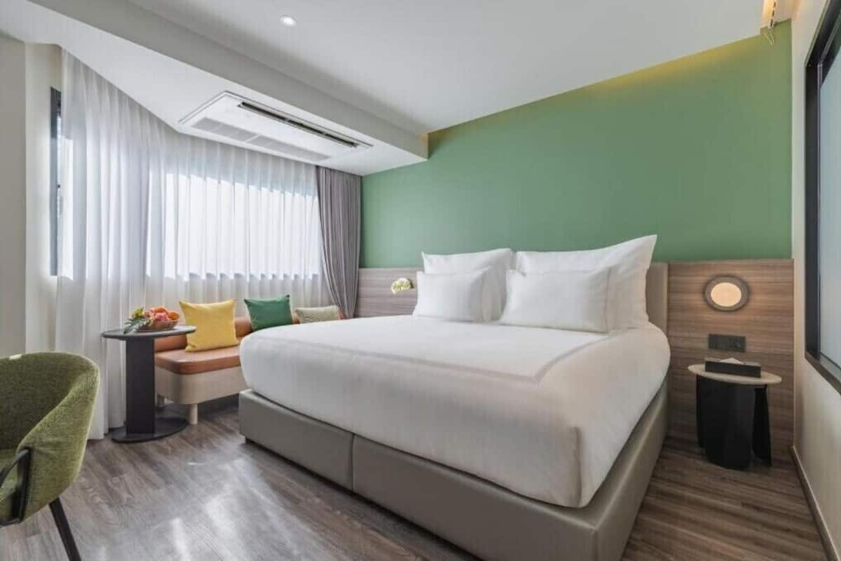 A hotel room with a bed, a side table, a chair, a window with curtains, and an air conditioning unit.