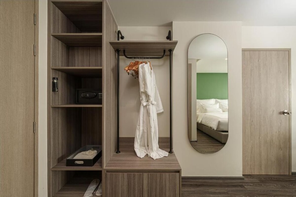 Habitación de hotel con un armario de madera, una bata colgada, un espejo y una cama frente a una pared verde.