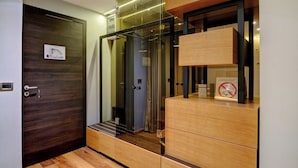 Une chambre d’hôtel moderne avec un plancher en bois, une porte en bois foncé, un ascenseur en verre et une armoire en bois dotée de tiroirs.