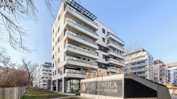Un edificio de varios pisos con balcones, un letrero que dice 'MIASTO WOLA' y un área verde al frente.
