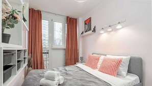 Ein ordentlich eingerichtetes Schlafzimmer mit einem Bett, einem Fenster mit Vorhängen, einem Regal mit Pflanzen und einer wandmontierten Leuchte.