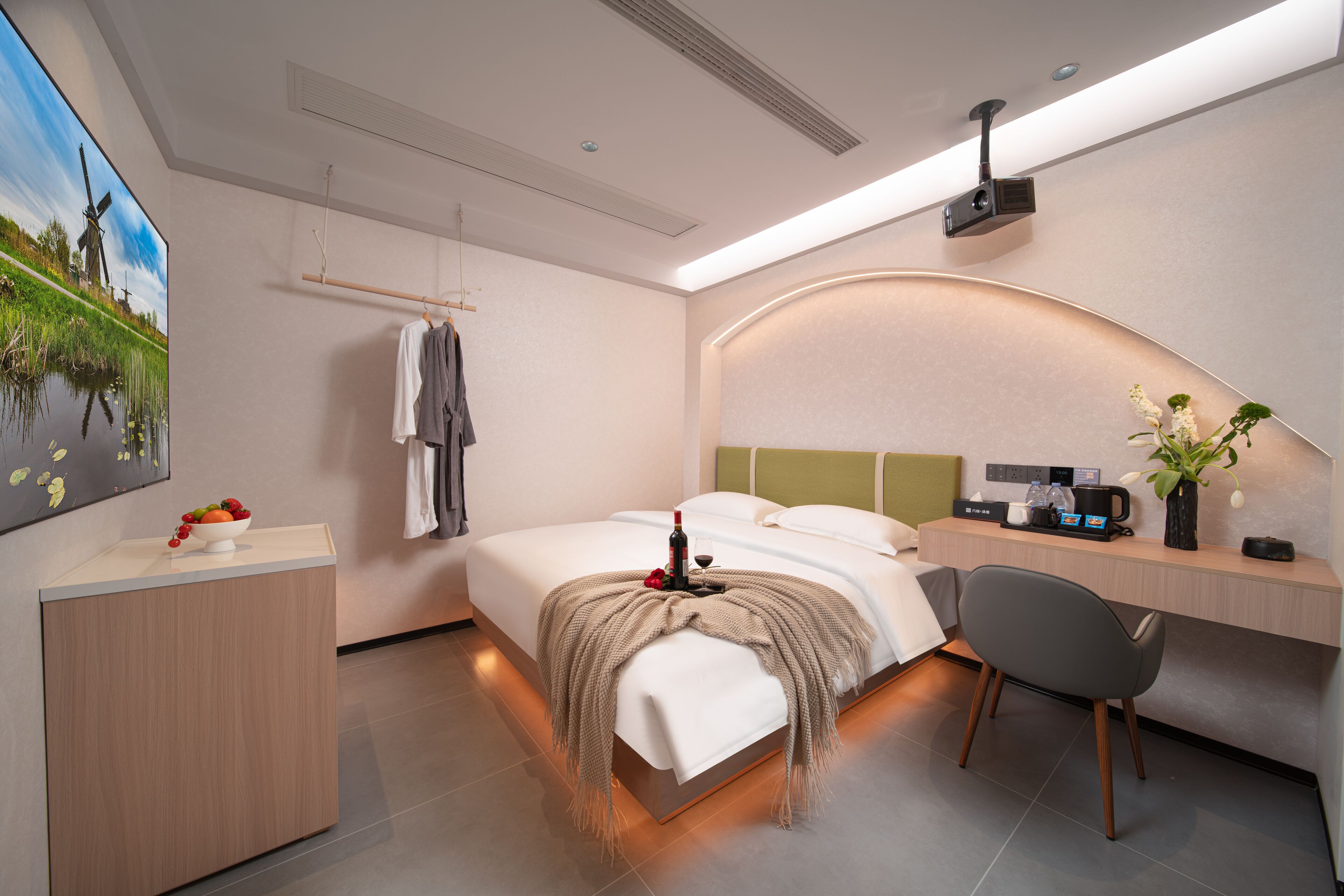 A modern hotel room with a bed, bedside table, desk, chair, and a painting on the wall.