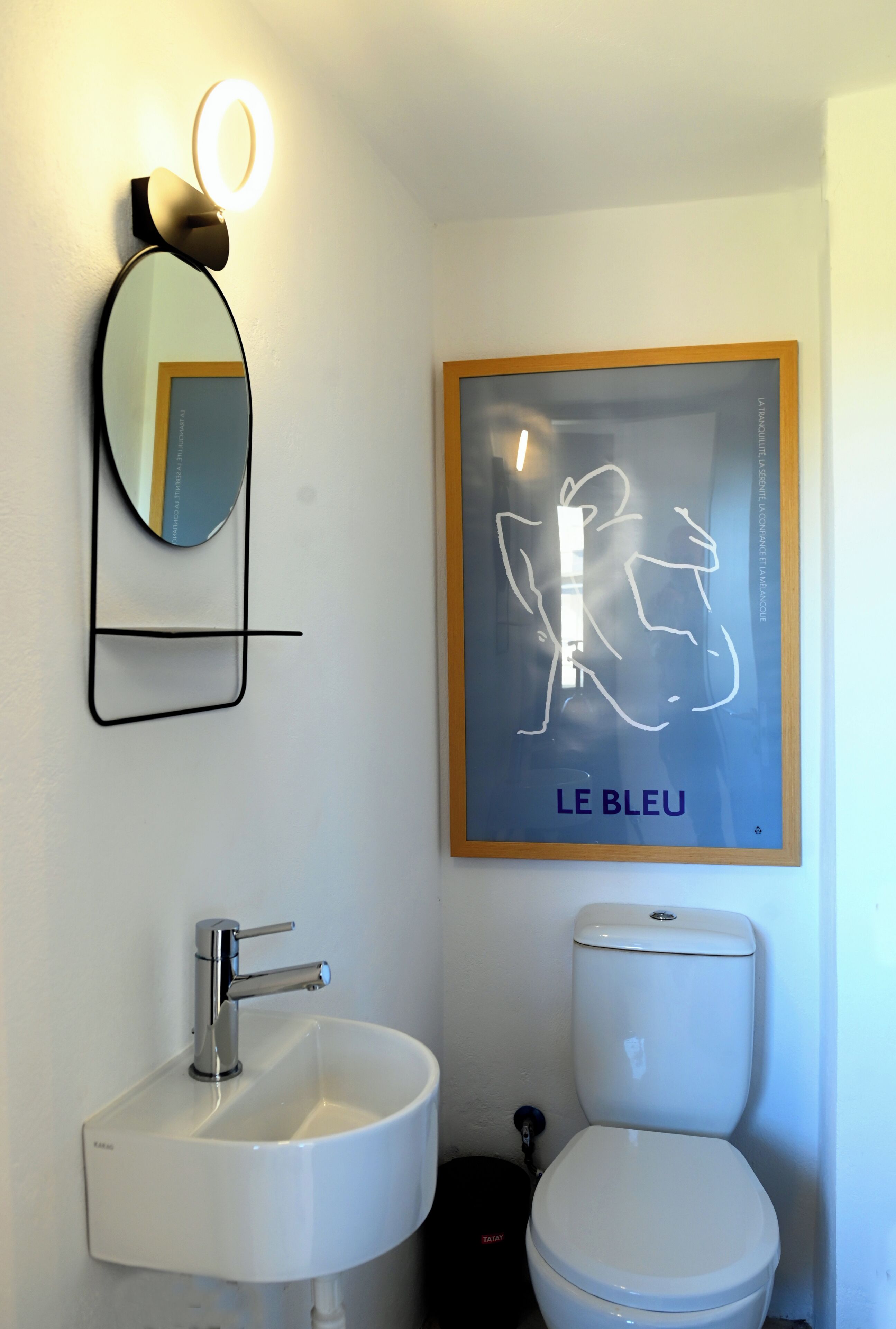 Ein Badezimmer mit einem weißen Waschbecken, einer Toilette, einem gerahmten abstrakten Kunstwerk und einem Spiegel mit Beleuchtung darüber.