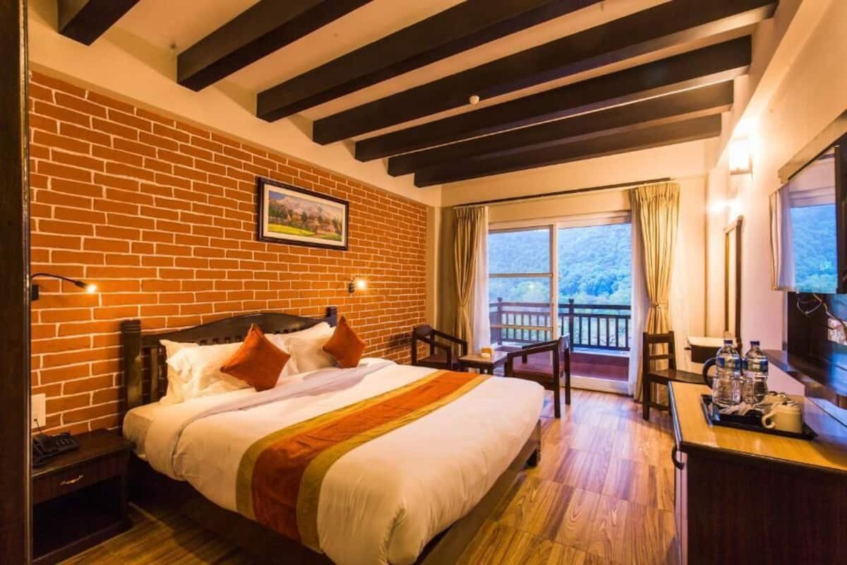 Una habitación de hotel con pared de ladrillo, cama con ropa blanca y detalles naranja, escritorio de madera con silla, y balcón con vistas.