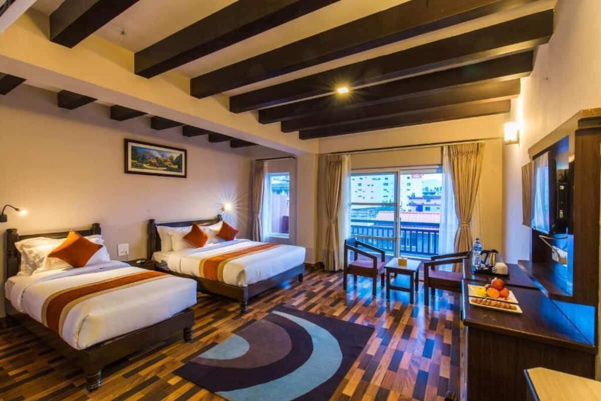 Habitación de hotel con dos camas, un balcón con vistas, un escritorio y televisor, y piso de madera.