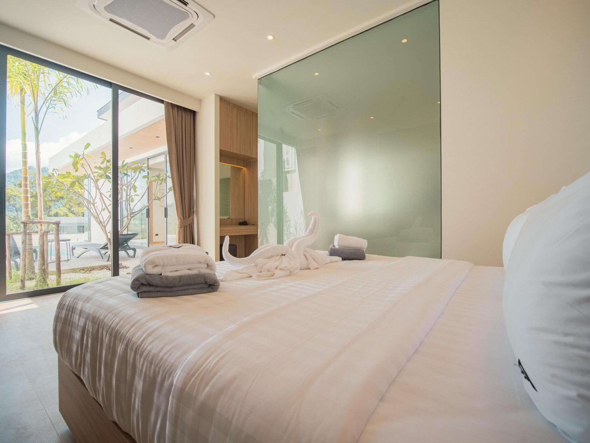 A modern hotel room with a large bed, a glass wall, and a view of the outdoors.