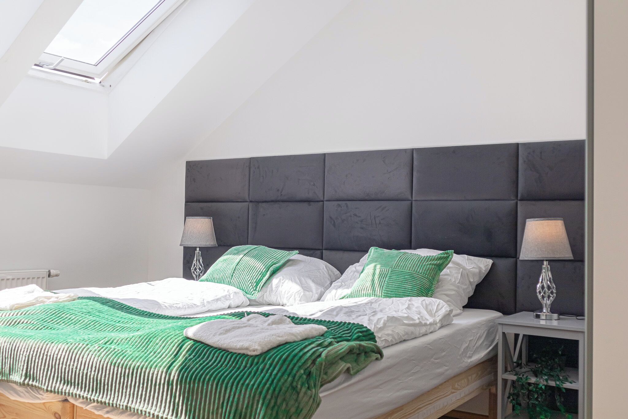 Een slaapkamer met een schuine kap, een donker hoofdbord, twee bedlampen en een bed met wit- en groen beddengoed.