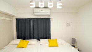 Een tweepersoonsbed met witte beddengoed en twee gele kussens, een donker gordijn, een wandairconditioner en een nachtkastje met een lamp.