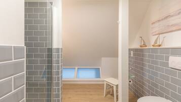 Ein Badezimmer mit grauen Fliesenwänden, einer weißen Toilette, einem kleinen weißen Stuhl und einem Fenster mit Blick auf den Himmel.