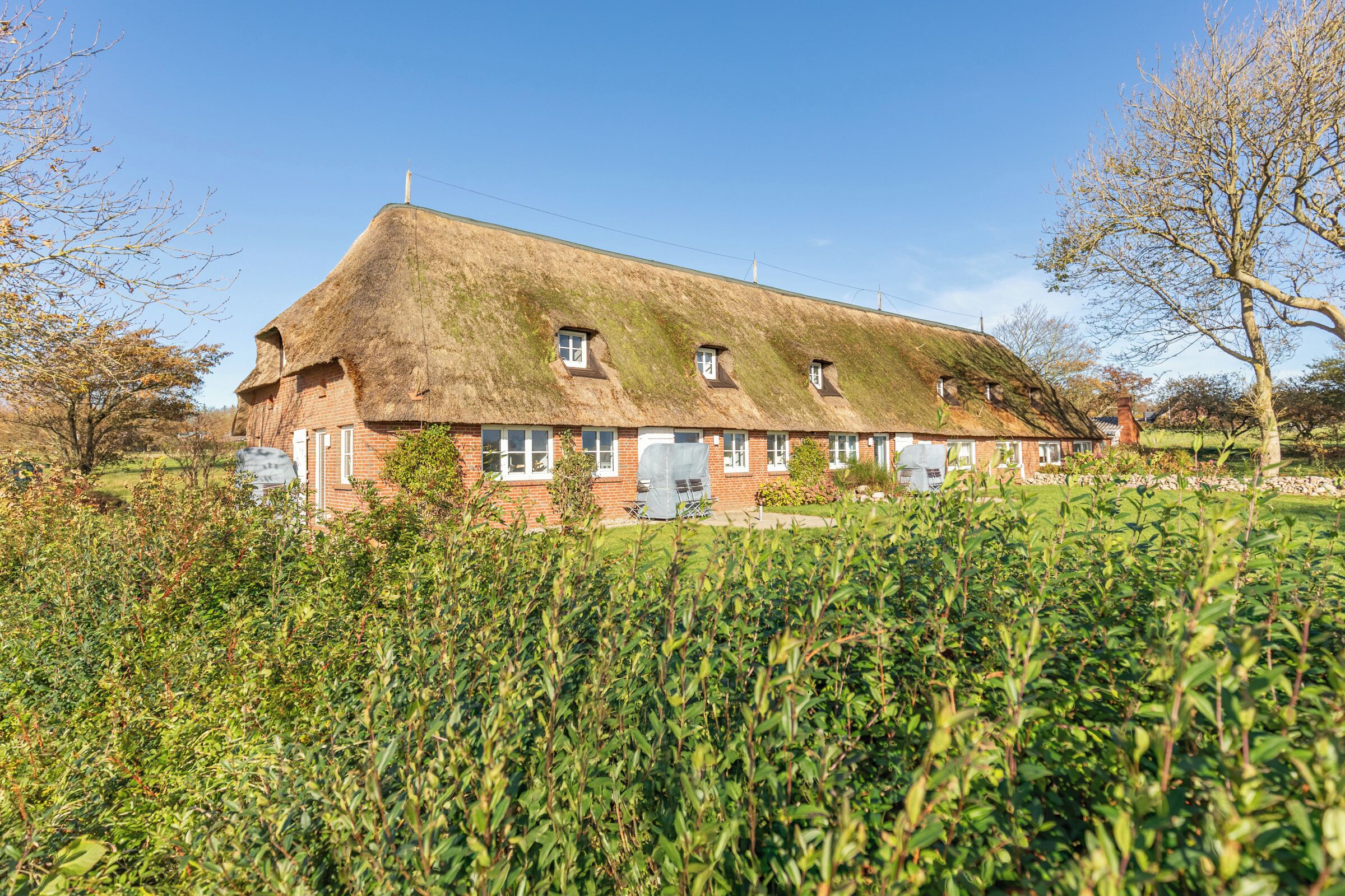 Ein traditionelles Haus mit Strohdach, zahlreichen Fenstern und Schornstein, umgeben von einem gepflegten Garten.