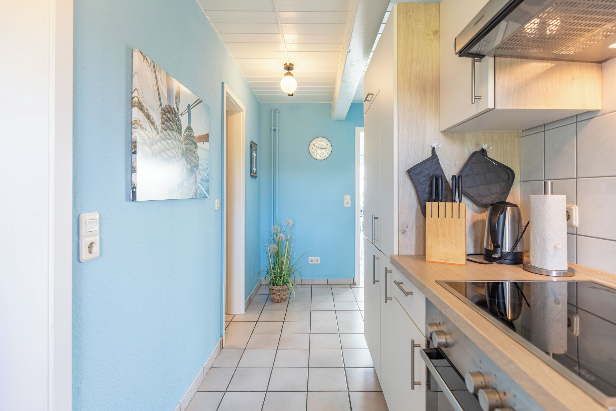 A narrow kitchen with light blue walls, a white ceiling, and a tiled floor. There is a clock on the wall, a plant in the corner, and a stove with a kettle on the counter.