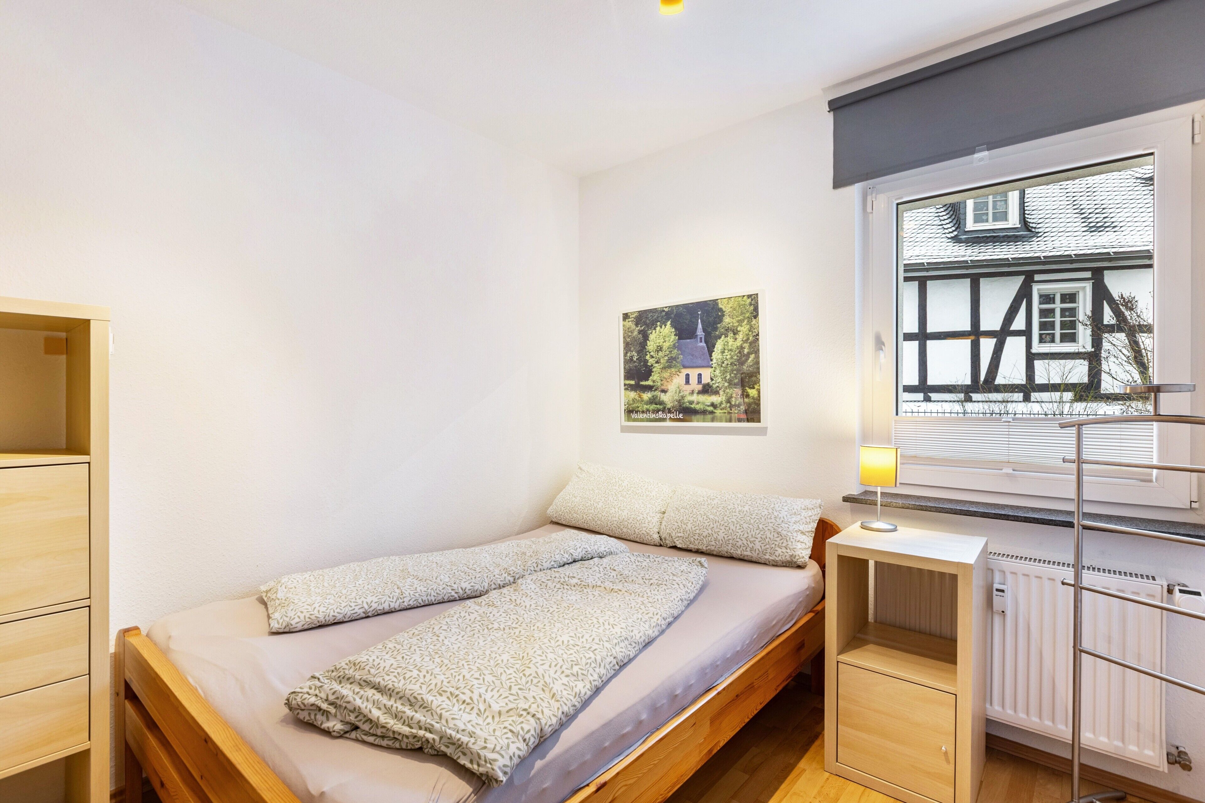 A single bed with a wooden frame, a bedside table, a radiator, and a window with a view of a house.