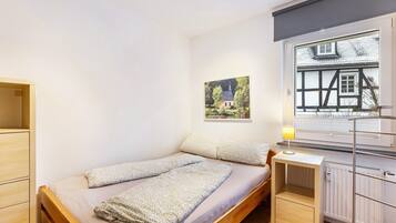 A single bed with a wooden frame, a bedside table, a radiator, and a window with a view of a house.