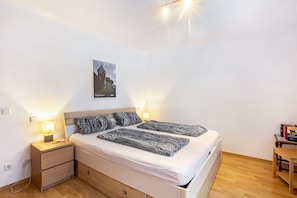 Un lit double avec une tête de lit, deux tables de chevet avec des lampes, un plancher en bois, un tableau accroché au mur et une petite table avec des livres.