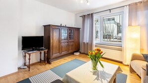 Een woonkamer met een televisie, een houten kast, een tafel met bloemen en een raam met gordijnen.