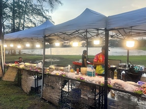 A food stall with a grill, ice, and various food items, set up under a canopy with a lake in the background. - Laguna Lake View Condo with Rooftop Pool – Skypark Bang Tao (ตำบลเชิงทะเล)
