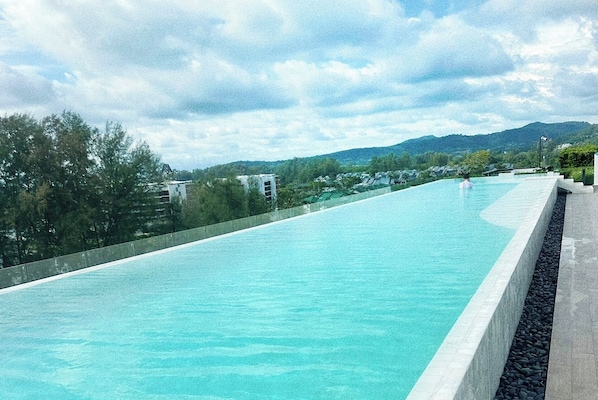 A long, rectangular swimming pool with clear blue water, surrounded by greenery and buildings in the background. - Laguna Lake View Condo with Rooftop Pool – Skypark Bang Tao (ตำบลเชิงทะเล)