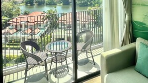 A balcony with a view of a lake, trees, and buildings. There are two wicker chairs and a small round table. - Laguna Lake View Condo with Rooftop Pool – Skypark Bang Tao (ตำบลเชิงทะเล)