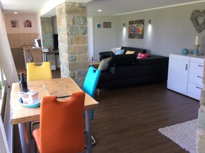 Un coin repas avec une table en bois, des chaises et une bouteille de vin. On aperçoit un canapé noir et un meuble blanc à l’arrière-plan.