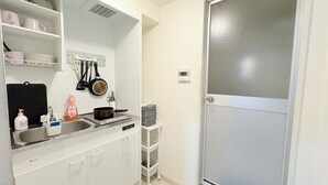 Una cucina compatta con mobili bianchi, un lavello e un piano cottura. Si vedono scaffali con stoviglie e un porta asciugamani. Accanto alla porta, un piccolo ripiano bianco con asciugamani piegati.