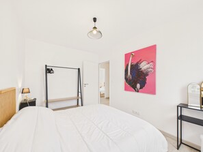 Ein Schlafzimmer mit einem Bett, einer Tür, einem Kleiderständer, einer Lampe, einem Ostrichgemälde und einer Deckenleuchte.