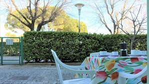 Un área de comedor al aire libre con una mesa preparada para dos personas, rodeada de sillas de plástico blanco, un mantel con estampado floral y una cafetera.
