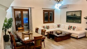 A dining area with a wooden table and chairs, a sofa with tropical pillows, and a painting on the wall. - Villa Logas Hill with Panoramic Sea Views, Wi-Fi and Air Conditioning (Peroulades)