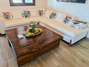 Una sala de estar con una mesa de centro de madera, sofá blanco con almohadones de temática tropical y una bandeja de frutas.
