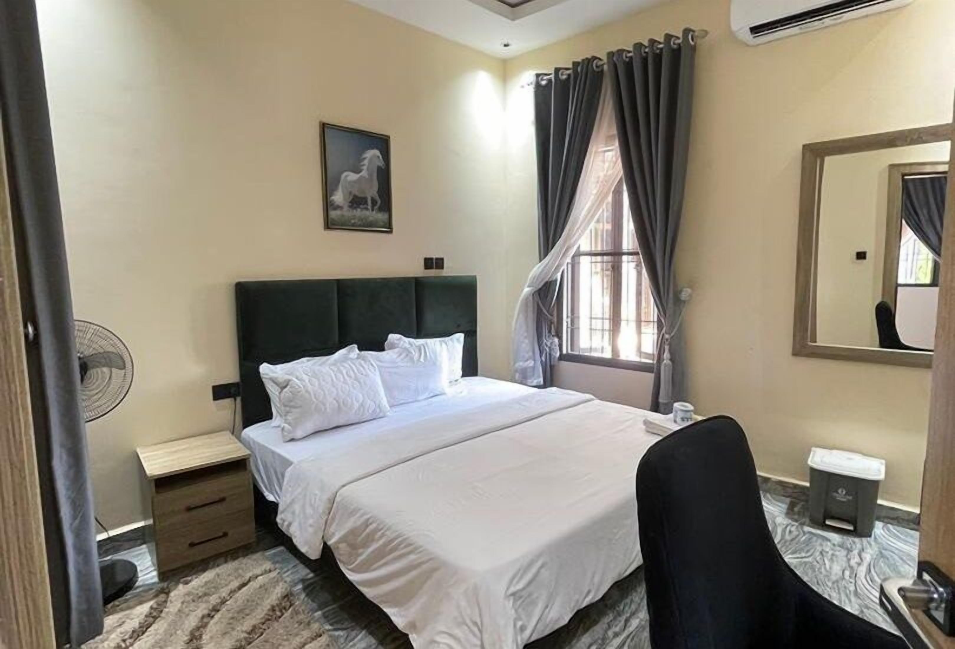 A hotel room with a bed, a nightstand, a chair, a fan, a window with curtains, and a mirror.