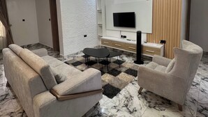 A modern living room with a sofa, armchair, and a TV on a wooden panel wall. - Deroche Apartment (Abuja)