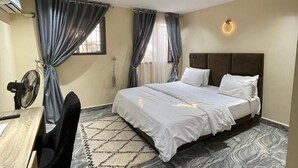 A hotel room with a bed, a desk, a chair, and a fan. - Deroche Apartment (Abuja)
