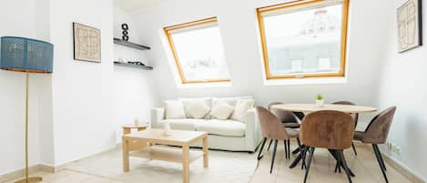 Una sala de estar moderna con un sofá, una mesa de centro y un comedor con sillas. Hay dos ventanas con vistas a edificios.