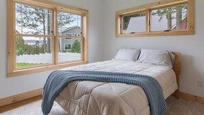 A bedroom with a bed, a window with a view of a house and trees, and a wooden headboard. - Cozy 2-bedroom guest house in perfect South Hero with private lake access. (South Hero)