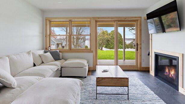 A modern living room with a sofa, a coffee table, a fireplace, and large windows offering a view of a green landscape. - Cozy 2-bedroom guest house in perfect South Hero with private lake access. (South Hero)