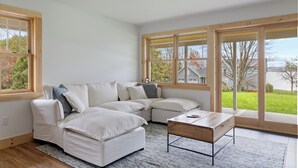 A modern living room with a sectional sofa, a coffee table, and large windows offering a view of the outdoors. - Cozy 2-bedroom guest house in perfect South Hero with private lake access. (South Hero)