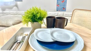 Una mesa de comedor con una bandeja blanca que contiene cubiertos, dos platos blancos con interiores azules, una pequeña planta en maceta y dos tazas oscuras.
