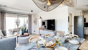 Een moderne woonkamer met een eethoek, gedekt voor een maaltijd, met een grote televisie, een bank en een eettafel met wijnglazen en brood.