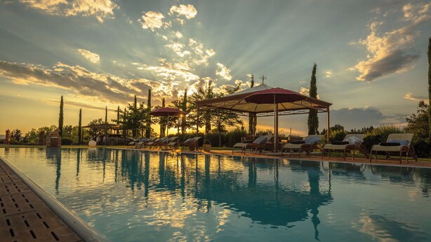 Area piscina con lettini e ombrelloni, immersa tra alberi e un gazebo.