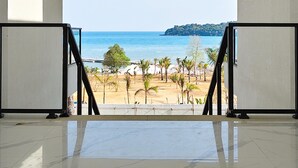A spacious room with a large window overlooking a beach and ocean, featuring a high ceiling and a tiled floor.