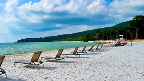 A beach with rows of sun loungers, a lifeguard tower, and a forested hill in the background.