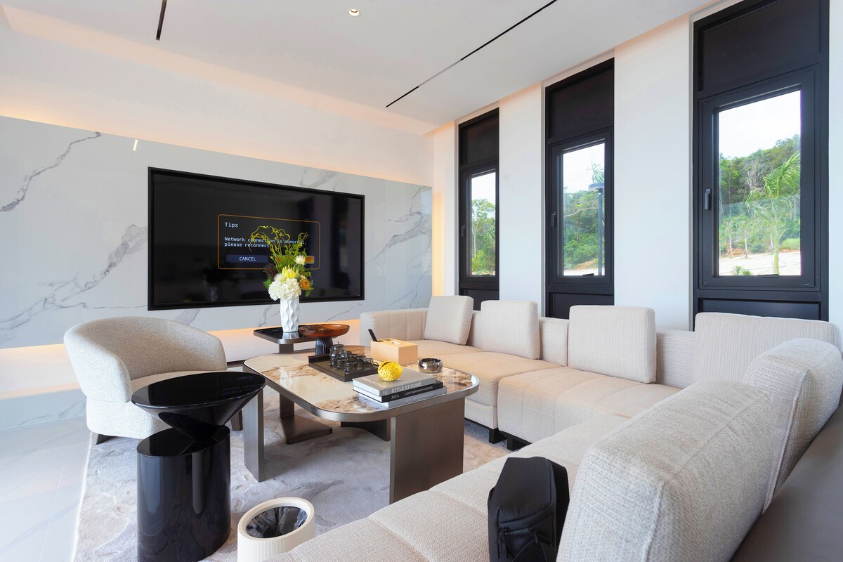 Una sala de estar moderna con un televisor de pantalla plana grande, un sofá color beige, una mesa de centro y un ventanal con vistas a zonas verdes.